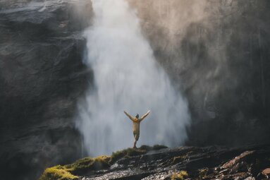 Person in a yellow jacket stands with arms outstretched in front of a roaring waterfall. | © Holiday Region National Park Hohe Tauern - Gerald Demolsky
