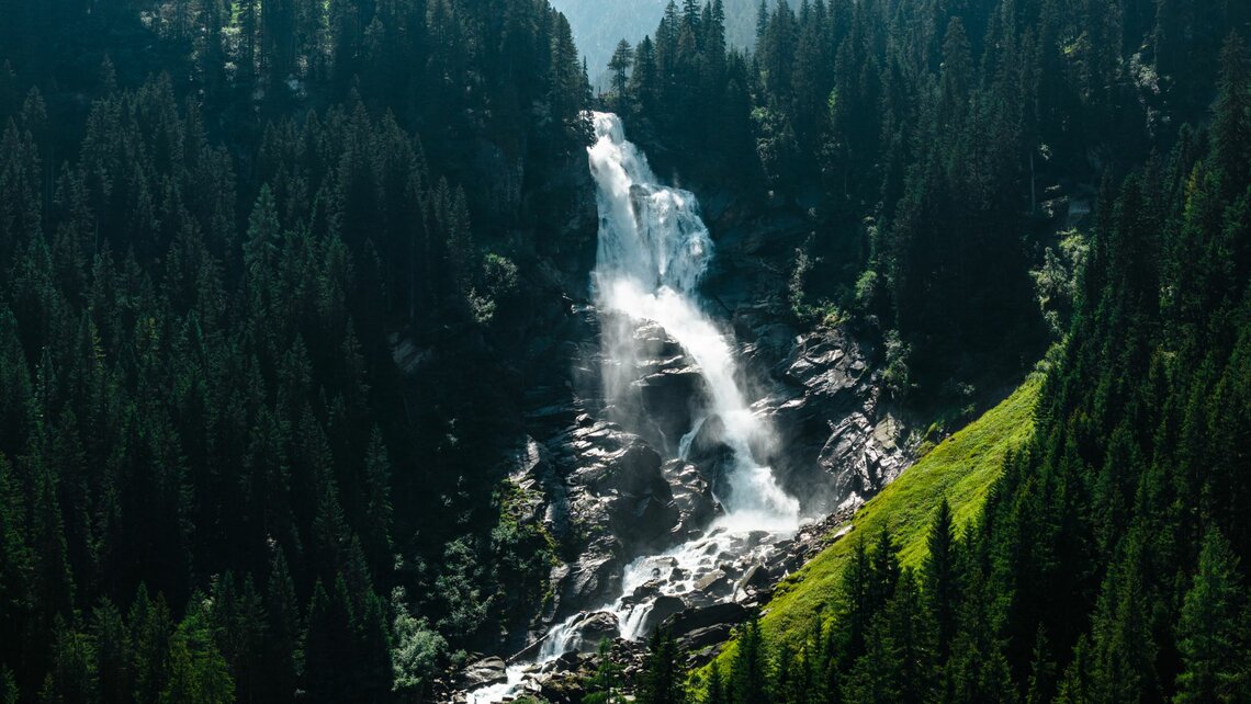 Mächtiger Wasserfall inmitten eines grünen Waldes mit Gischt und Sonnenlicht. | © Ferienregion NAtionalpark Hohe Tauern - Mathäus Gartner