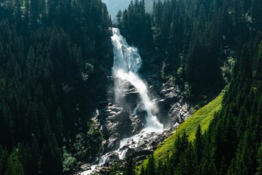 Mächtiger Wasserfall inmitten eines grünen Waldes mit Gischt und Sonnenlicht. | © Ferienregion NAtionalpark Hohe Tauern - Mathäus Gartner