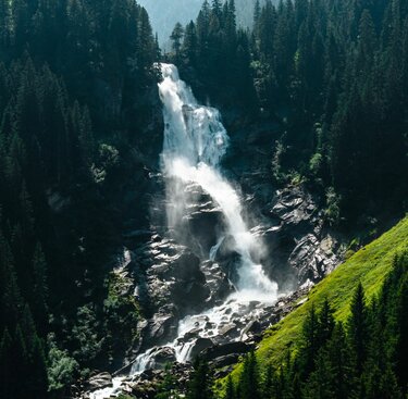 Mächtiger Wasserfall inmitten eines grünen Waldes mit Gischt und Sonnenlicht. | © Ferienregion NAtionalpark Hohe Tauern - Mathäus Gartner