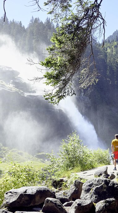 Paar steht auf einem Weg und blickt auf einen großen Wasserfall in sonniger Natur. | © Ferienregion Nationalpark Hohe Tauern - Michael Huber