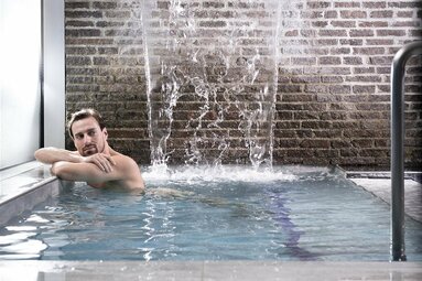 Mann entspannt in einem Pool mit Wasserfall vor einer Backsteinwand in moderner Umgebung. | © Gasteinertal Tourism GmbH - Marktl