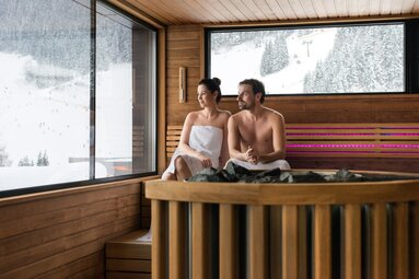 Paar in einer Sauna mit Panoramafenster und Blick auf verschneite Berge und Skipisten. | © Gasteinertal Tourismus GmbH - Marktl Photography