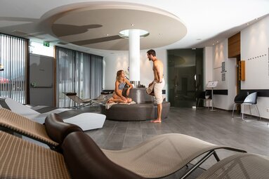 Entspannungsbereich in einem Spa mit Liegen, moderner Einrichtung und zwei Personen im Gespräch. | © Kristallbad Wald - Königsleiten