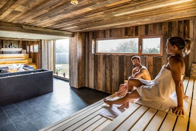 Paar entspannt in einer gemütlichen Holzsauna mit großem Fenster und moderner Einrichtung. | © Ferienregion Nationalpark Hohe Tauern