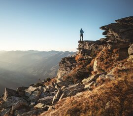 Mann steht auf einem Felsvorsprung und blickt in eine weite Berglandschaft bei Sonnenaufgang.  | © Ferienregion Nationalpark Hohe Tauern - Daniel Breuer