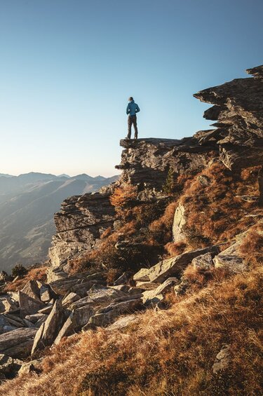 Man stands on a rocky outcrop and looks out over a vast mountain landscape at sunrise. | © Holiday Region National Park Hohe Tauern - Daniel Breuer