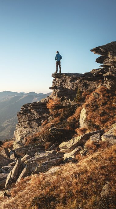 Mann steht auf einem Felsvorsprung und blickt in eine weite Berglandschaft bei Sonnenaufgang.  | © Ferienregion Nationalpark Hohe Tauern - Daniel Breuer
