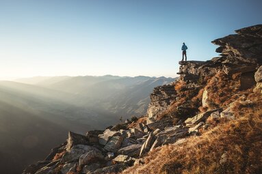 Mann steht auf einem Felsvorsprung und blickt in eine weite Berglandschaft bei Sonnenaufgang.  | © Ferienregion Nationalpark Hohe Tauern - Daniel Breuer