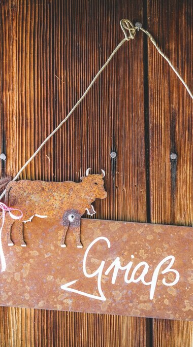 Rostiges Metallschild mit Kuhfigur und der Aufschrift „Griaß di!“ an einer Holzwand. | © Ferienregion Nationalpark Hohe Tauern