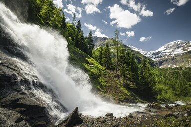 Wasserfall in den Alpen, umgeben von grünen Wäldern und schneebedeckten Bergen unter blauem Himmel. | © Ferienregion Nationalpark Hohe Tauern - Max Draeger