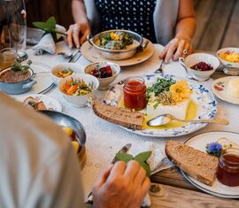 Rustikale Brotjause mit Käse, Brot, Kräutern und Beilagen auf einem Holztisch, zwei Personen essen. | © SalzburgerLand Tourismus - Chris Perkles