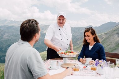 Koch serviert Kaiserschmarrn an zwei Gäste auf einer Terrasse mit Bergblick. | © SalzburgerLand Tourismus - Chris Perkles