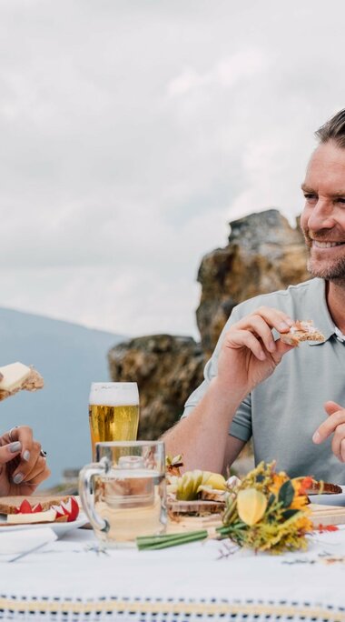 A smiling couple enjoys a meal with snacks and beer at a table in the mountains. | © SalzburgerLand Tourism - Chris Perkles