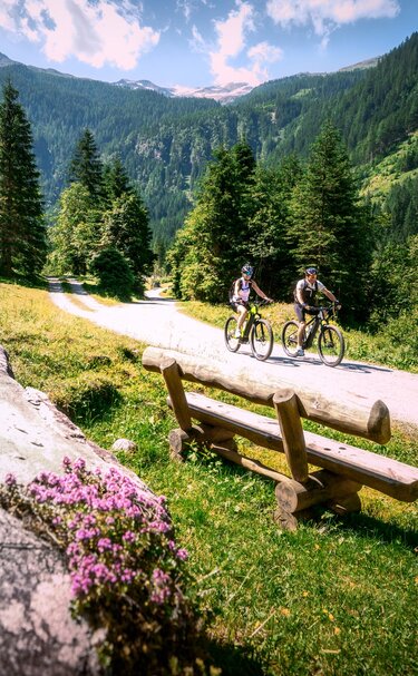 Zwei Mountainbiker fahren an Rastbank auf Almweg vorbei. | © Daniel Kogler