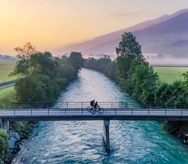 Zwei Radfahrer überqueren eine Brücke über einen ruhigen Fluss, umgeben von Wiesen, Bäumen und sanften Bergen im frühen Morgenlicht. | © Lichtfarben