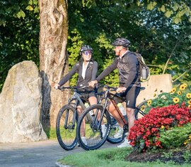 Zwei Radfahrer halten auf einem Weg neben buntem Blumenbeet im Grünen. | © Lichtfarben