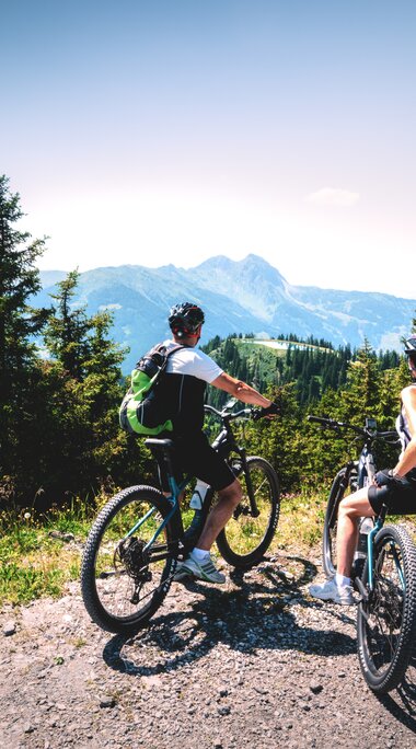 Two mountain bikers resting on a gravel path with alpine panorama. | © Hohe Tauern National Park holiday region - Daniel Kogler