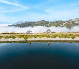 Zwei Radfahrer fahren entlang eines Bergsees über den Wolken mit Spiegelung im Wasser | © Wildkogel-Arena Neukirchen & Bramberg
