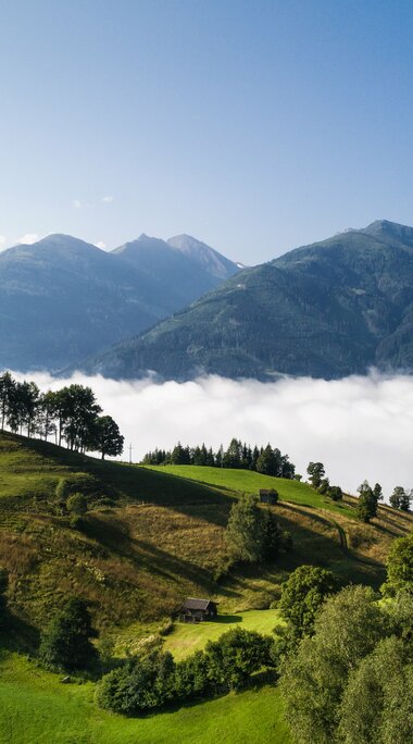 Grüne Hügel unter klarem Himmel, mit Nebel in einem Tal und Bergen im Hintergrund. | © Ferienregion Nationalpark Hohe Tauern - David Innerhofer