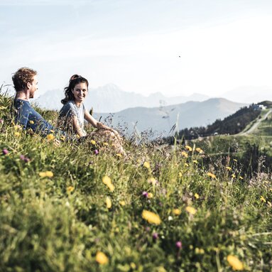 Couple sitting in a flower meadow with mountain panorama in the background, enjoying the view. | © Zell am See-Kaprun Tourism