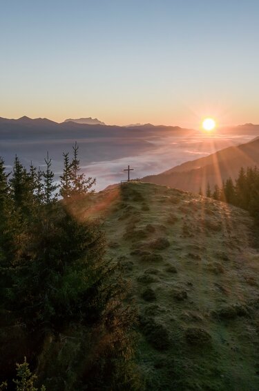 Sonnenaufgang über Bergen mit einem Gipfelkreuz und Bäumen im Vordergrund. | © Mittersill plus Tourismus