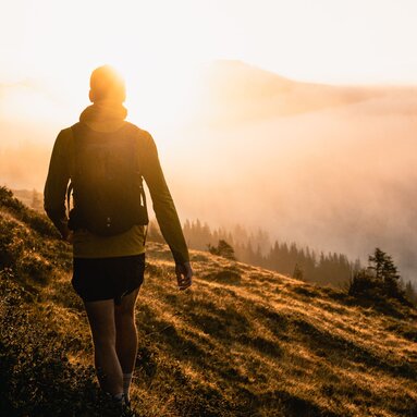 Eine Person wandert bei Sonnenaufgang auf einem grasbewachsenen Hügel mit Blick auf nebelbedeckte Wälder. | © Ferienregion Nationalpark Hohe Tauern - Gerald Demolsky