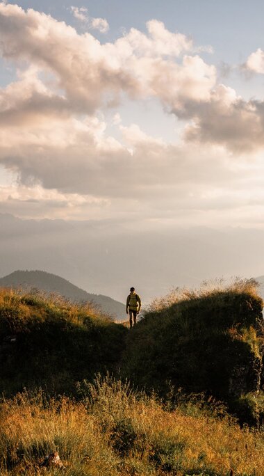 Eine Person steht auf einem grasbewachsenen Hügel mit weitem Blick auf Berge und Wolken im Sonnenlicht. | © Ferienregion Nationalpark Hohe Tauern - Gerald Demolsky
