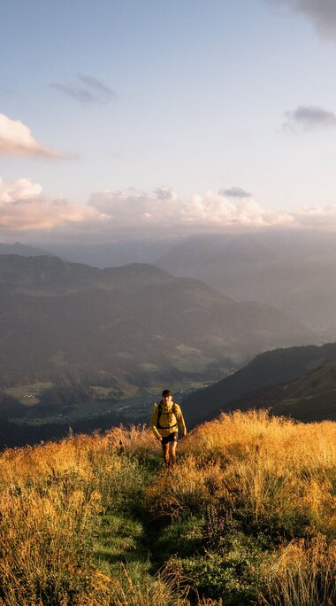 Eine Person wandert auf einem goldenen Hügelpfad mit Blick auf eine Berglandschaft bei Sonnenuntergang. | © Ferienregion Nationalpark Hohe Tauern - Gerald Demolsky