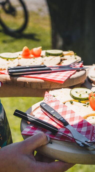 Eine Person serviert auf Holzbrett angerichtete Brotjause mit Käse, Gurken und Tomaten im Freien. | © Ferienregion Nationalpark Hohe Tauern - Gerald Demolsky