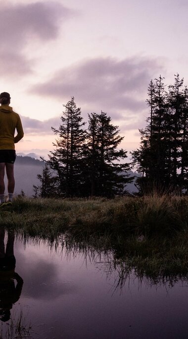 A person stands at dusk by a reflective pond, surrounded by trees and mist. | © Holiday Region National Park Hohe Tauern - Gerald Demolsky
