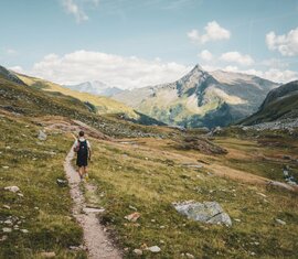 Hiker on a mountain trail, surrounded by meadows and peaks under a cloudy sky. | © Holiday Region National Park Hohe Tauern - Gerald Demolsky