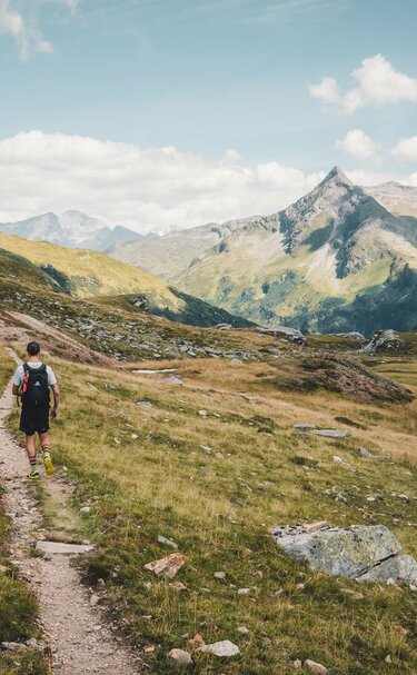 Wanderer auf einem Bergpfad, umgeben von Wiesen und Gipfeln unter einem bewölkten Himmel. | © Ferienregion Nationalpark Hohe Tauern - Gerald Demolsky