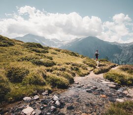 Wanderer auf einem Gebirgspfad, umgeben von Wiesen und Bergen unter einem bewölkten Himmel. | © Ferienregion Nationalpark Hohe Tauern - Gerald Demolsky