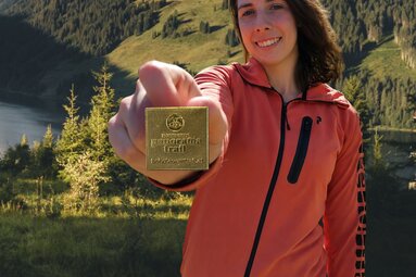 Wanderin zeigt die goldene Wandernadel vor Bergsee in den Hohen Tauern. | © Daniel Kogler - KI-unterstützt