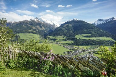 View of a green valley with a village, surrounded by snow-capped mountains and a wooden fence. | © Tourismusverband Piesendorf Niedernsill - Harry Liebmann