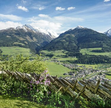 Blick auf ein grünes Tal mit einem Dorf, umgeben von schneebedeckten Bergen und einem Holzzaun. | © Tourismusverband Piesendorf Niedernsill - Harry Liebmann
