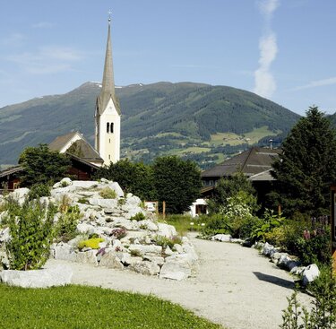 Ein Garten, umgeben von Bergen und blühender Landschaft, im Hintergrund ist die Kirche zu  sehen. | © Michael Huber