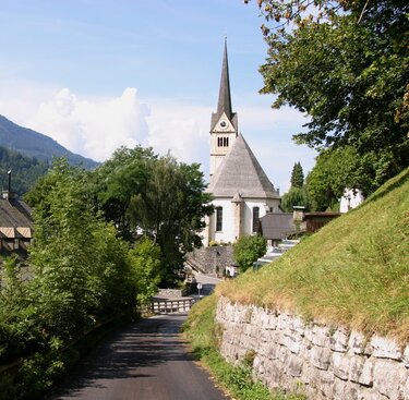 Dorfstraße mit Bäumen, Häusern und einer Kirche mit spitzem Turm, umgeben von Bergen. | © Tourismusverband Taxenbach