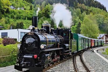 Historische Dampflokomotive auf Schienen, umgeben von grünen Bäumen und Bergen im Hintergrund. | © Pinzgauer Lokalbahn