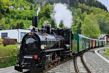 Historische Dampflokomotive auf Schienen, umgeben von grünen Bäumen und Bergen im Hintergrund. | © Pinzgauer Lokalbahn