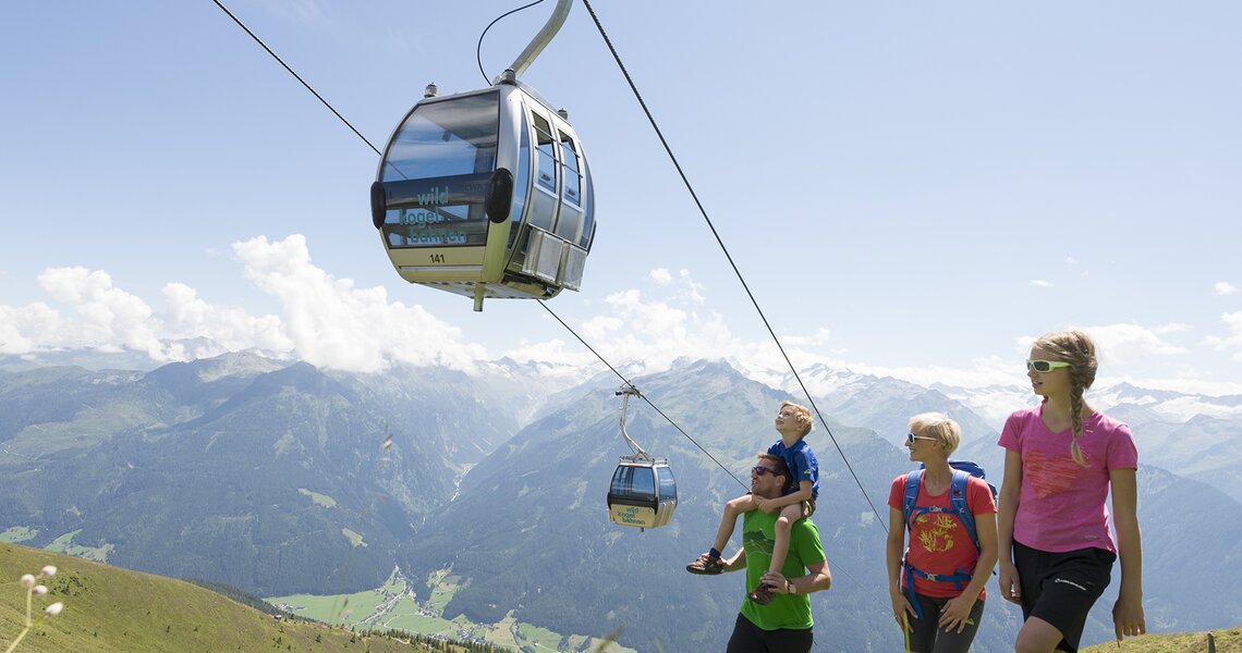 Familie wandert unter einer Gondelbahn der Wildkogelbahnen, Berge im Hintergrund. | © Bergbahnen Wildkogel - Wildkogelbahn Neukirchen