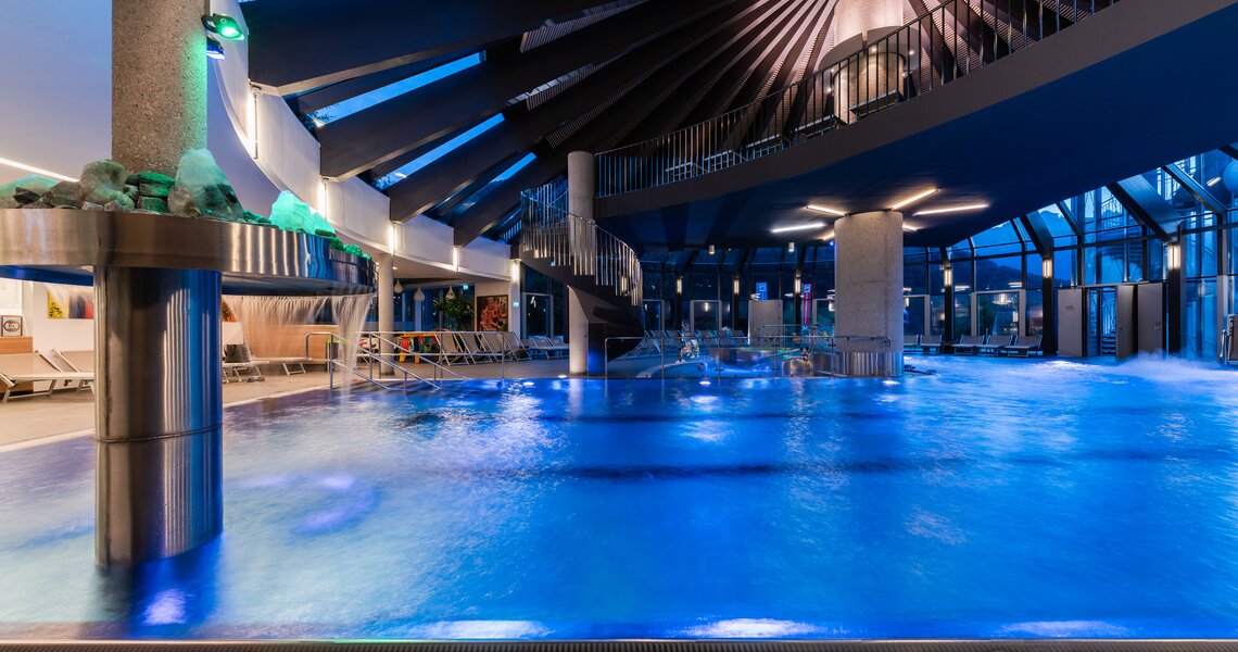 Innenansicht eines modernen Hallenbades mit blau beleuchtetem Wasser und Glaskuppel. | © Kristallbad Wald Königsleiten