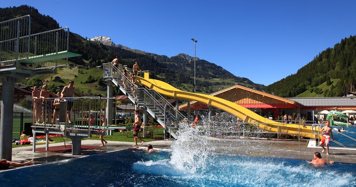 Freibad mit gelber Wasserrutsche, Sprungturm und Besuchern, umgeben von Bergen und grüner Natur. | © Freibad Großarl