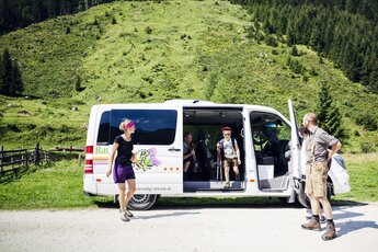 Wanderer steigen aus einem Nationalparktaxi in grüner Almlandschaft aus, umgeben von Wäldern. | © Seidlwinkl Nationalpark Taxi