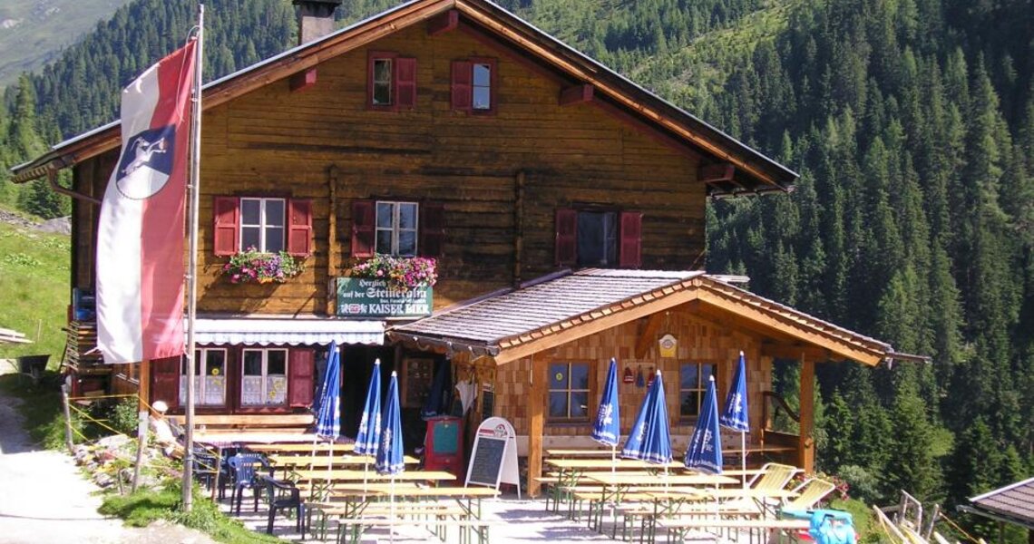 Traditionelle Berghütte mit Holzbalkon und Terrasse inmitten von grünen Wiesen und Wäldern. | © Posch Reisen