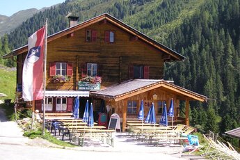 Traditionelle Berghütte mit Holzbalkon und Terrasse inmitten von grünen Wiesen und Wäldern. | © Posch Reisen