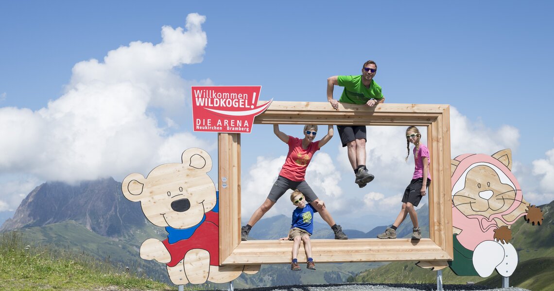 Familie posiert in einem Holzrahmen am Wildkogel, mit Blick auf Berge und Himmel. | © Bergbahnen Wildkogel - Smaragdbahn Bramberg