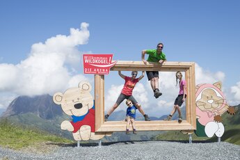 Familie posiert in einem Holzrahmen am Wildkogel, mit Blick auf Berge und Himmel. | © Bergbahnen Wildkogel - Smaragdbahn Bramberg