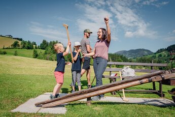 Familie spielt begeistert auf einer Holzrampe inmitten einer malerischen Wiesenlandschaft. | © Sobietzki